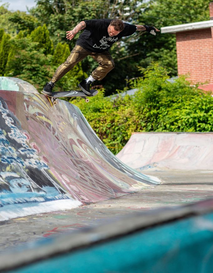 A skateboarder in a black graphic t-shirt and metallic gold pants performs a trick on a graffiti-covered ramp at an outdoor skatepark, with green trees and a brick building in the background.